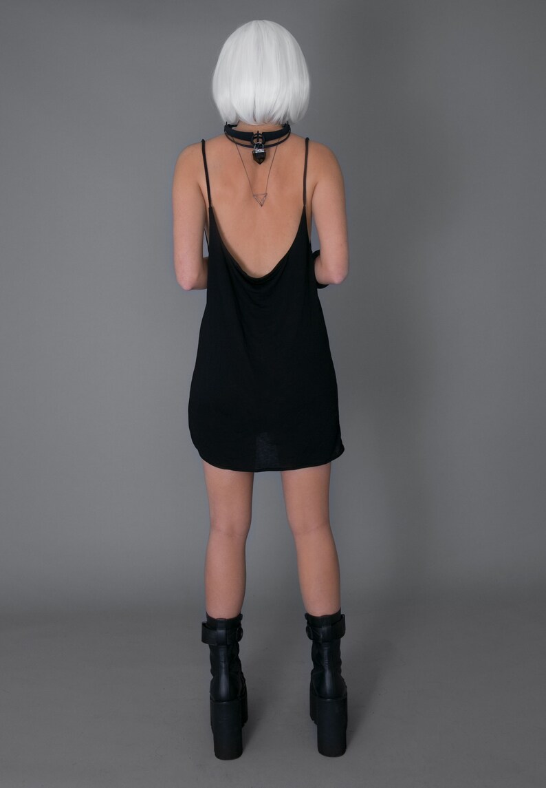 May include: A woman wearing a black sleeveless dress with a low back and black platform boots. She is wearing a silver necklace with a pendant.