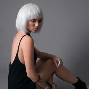 May include: A woman with short, white hair is wearing a black sleeveless dress and silver rings. She is sitting on a gray background with her legs crossed and wearing black sandals.