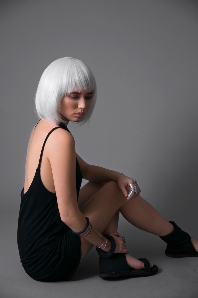 May include: A woman with short, white hair is wearing a black sleeveless dress and silver rings. She is sitting on a gray background with her legs crossed and wearing black sandals.