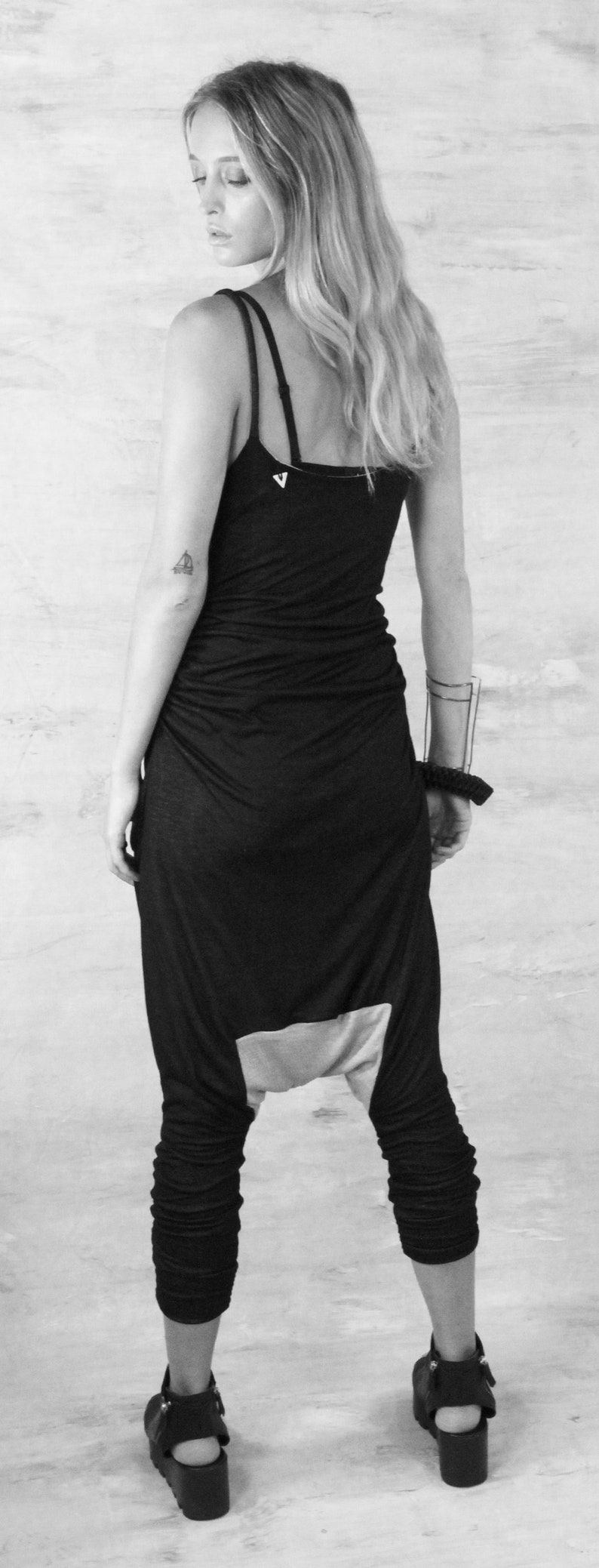 May include: A black and white photo of a person wearing a black jumpsuit with a loose fit and a low crotch. The jumpsuit has a white panel on the back. The person is wearing black platform sandals.