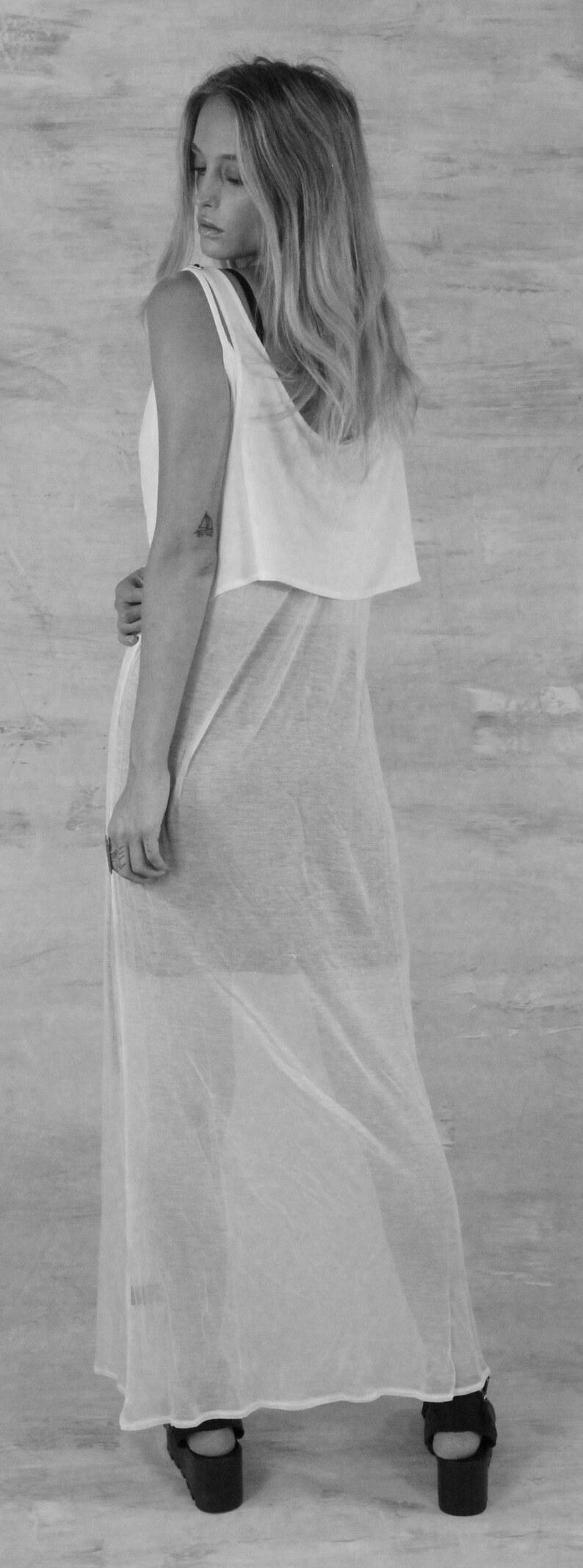 May include: A woman wearing a white sheer maxi dress with a white tank top underneath. She is wearing black platform sandals.