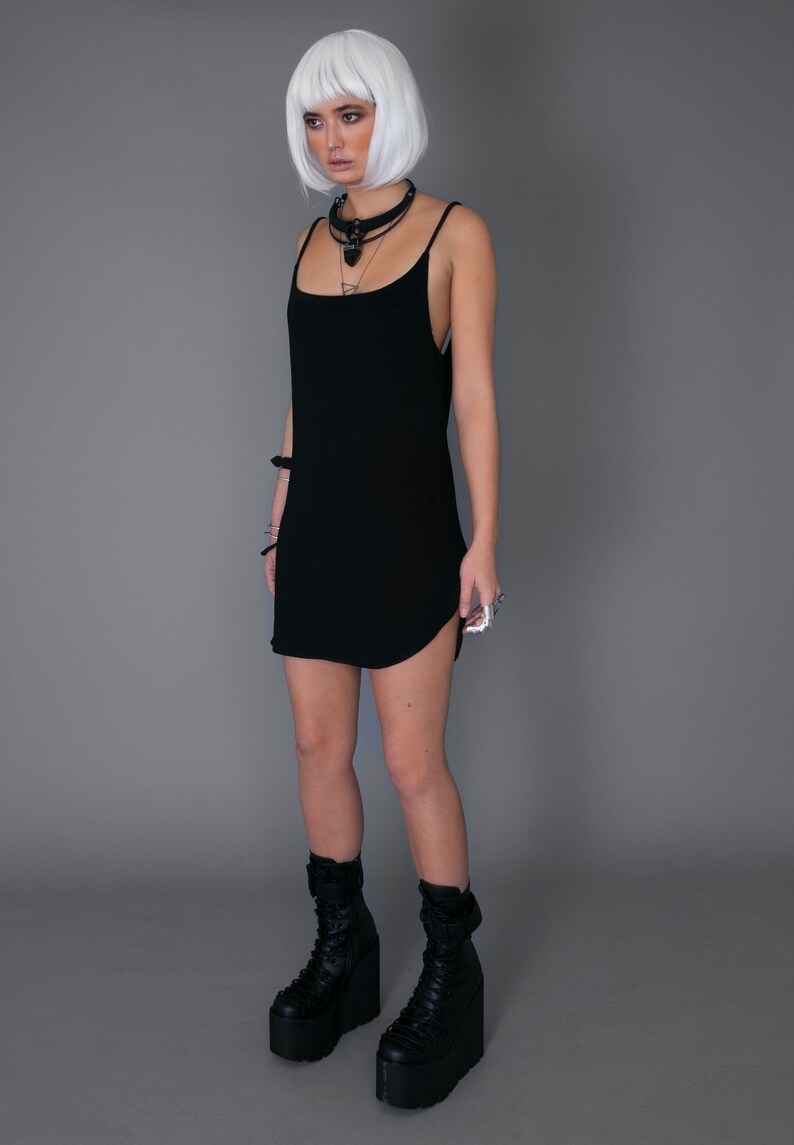 May include: A black sleeveless dress with a curved hemline, worn with black platform boots. The model has white hair and is wearing a black choker necklace.