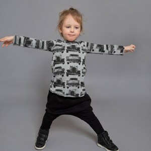 May include: A young child wearing a gray long-sleeved shirt with a black and white car print and black pants. The child is standing with their arms outstretched and wearing black shoes.