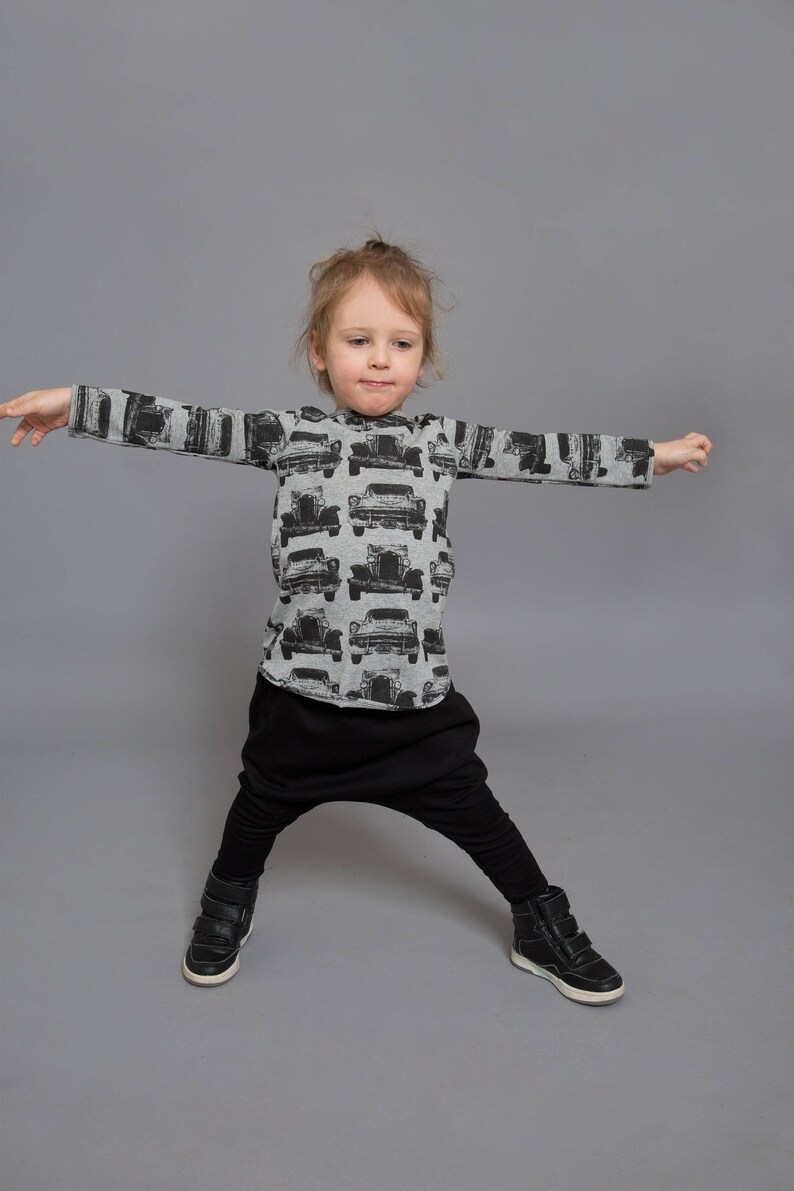 May include: A young child wearing a gray long-sleeved shirt with a black and white car print and black pants. The child is standing with their arms outstretched and wearing black shoes.