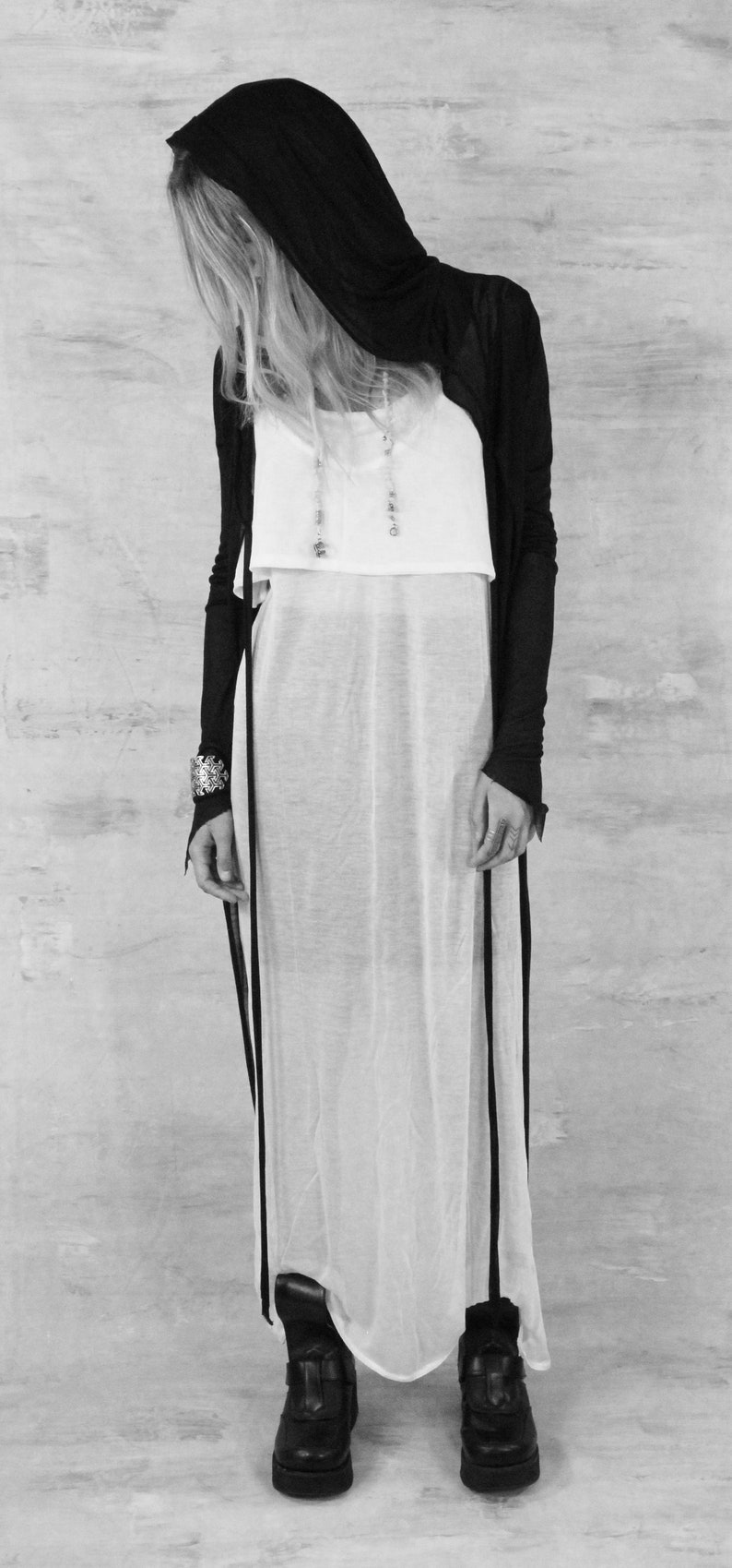 May include: A black hooded cardigan with long sleeves and a white sheer maxi dress. The cardigan has a long, flowing silhouette and the dress has a simple, elegant design.