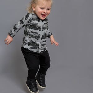 May include: A young child wearing a grey long-sleeved shirt with a black and white car print, black pants, and black high-top sneakers jumps in the air with a big smile.