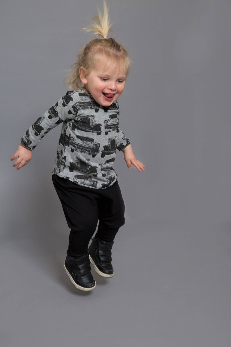 May include: A young child wearing a grey long-sleeved shirt with a black and white car print, black pants, and black high-top sneakers jumps in the air with a big smile.