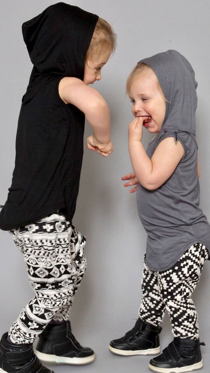 May include: Two children wearing hooded shirts and black and white patterned pants. The child on the left is wearing a black hooded shirt and the child on the right is wearing a gray hooded shirt.