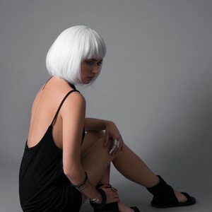 May include: A woman with short, white hair is wearing a black sleeveless dress and black sandals. She is sitting on a gray background with her legs crossed.