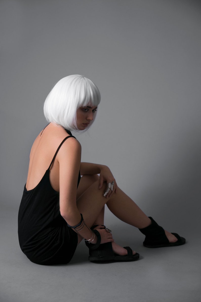 May include: A woman with short, white hair is wearing a black sleeveless dress and black sandals. She is sitting on a gray background with her legs crossed.