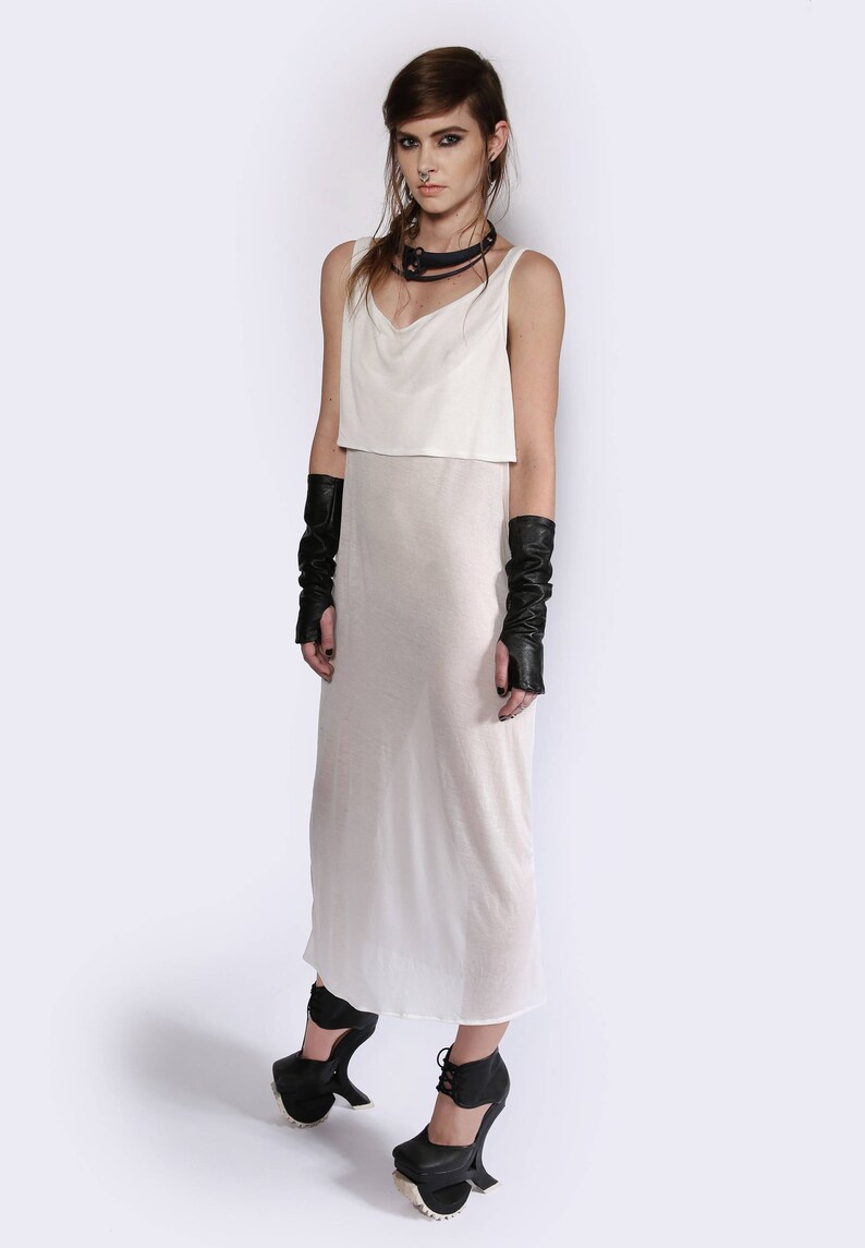 May include: A white sleeveless dress with a sheer overlay and a black choker necklace. The model is wearing black leather fingerless gloves and black platform shoes.