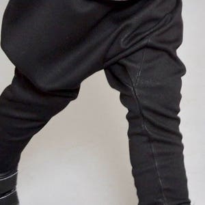 May include: Black pants with a gathered, loose fit around the thighs and a slim fit from the knees down. The pants are worn with black high-top sneakers.
