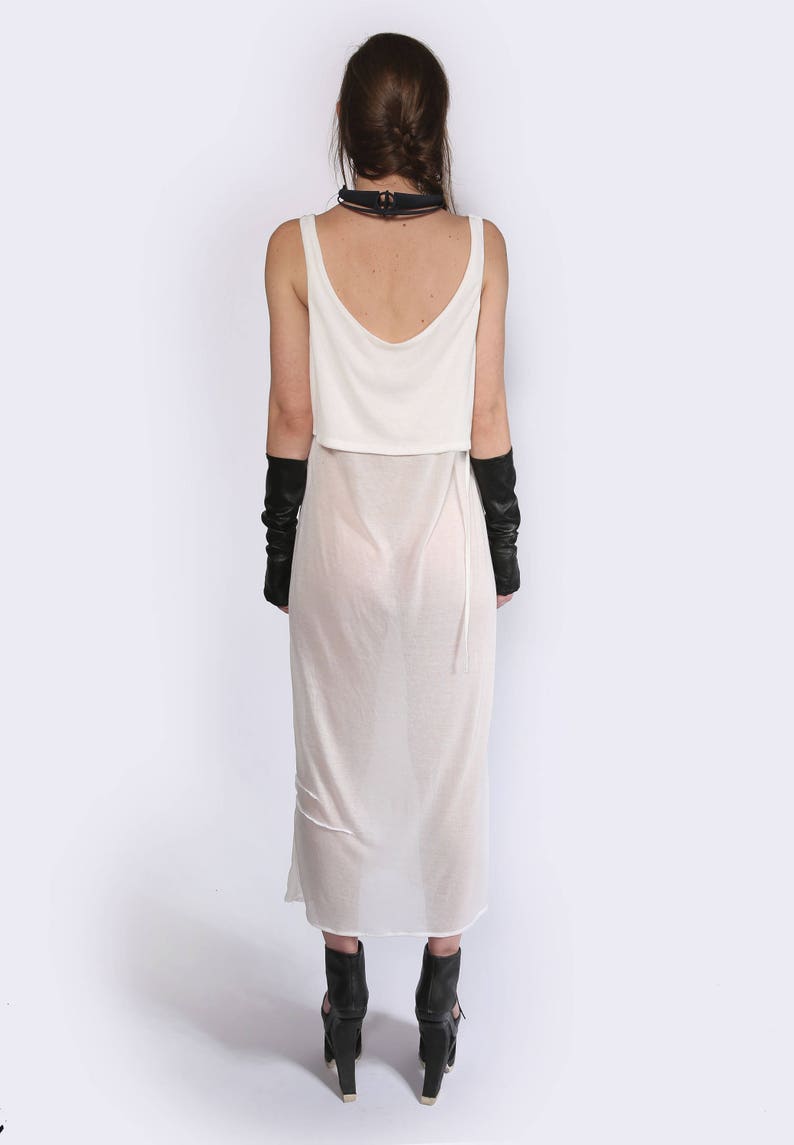 May include: A white sheer maxi dress with a white tank top layered over it. The dress has a thin black strap around the neck and the model is wearing black fingerless gloves.