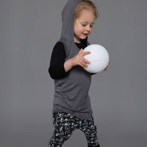 May include: A young child wearing a gray hooded shirt, black long-sleeved shirt, and black and white patterned pants, holds a white ball in their hands.