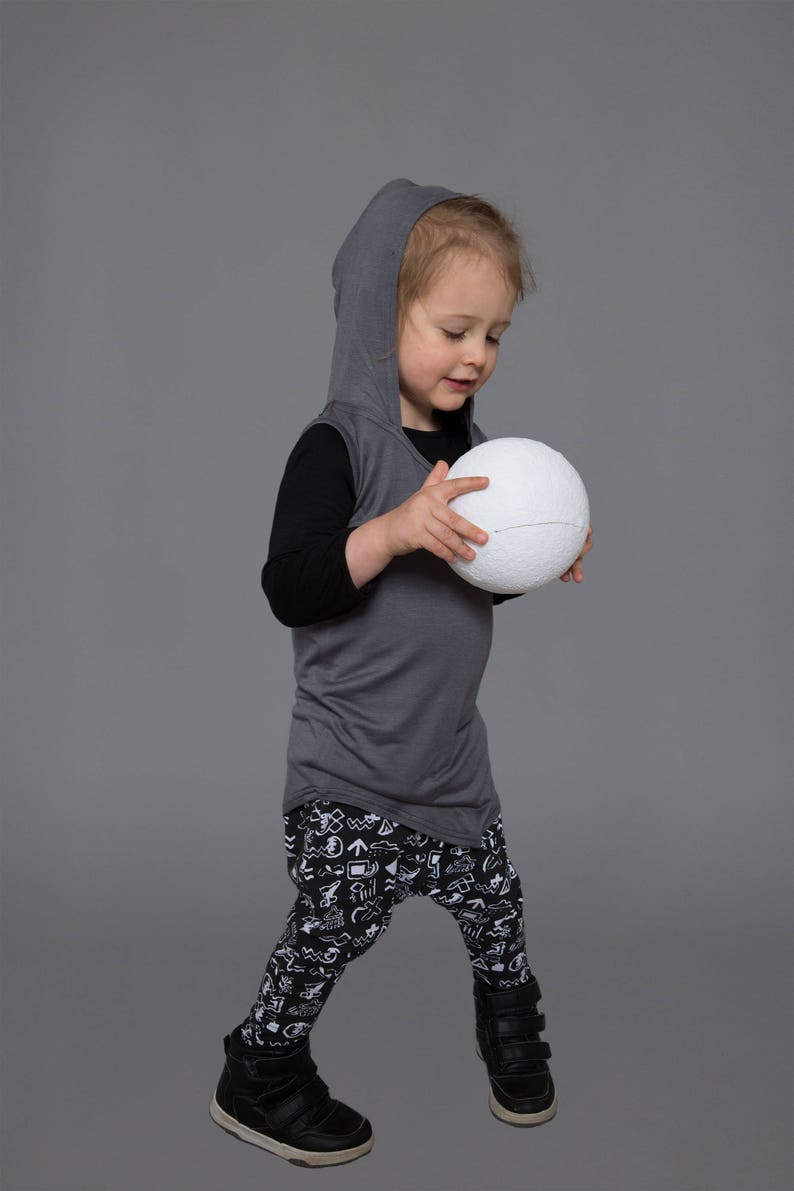 May include: A young child wearing a gray hooded shirt, black long-sleeved shirt, and black and white patterned pants, holds a white ball in their hands.