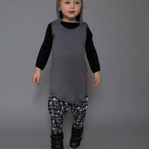 May include: A young person wearing a gray hooded shirt with black sleeves and black and white patterned pants. The person is standing on a gray background and looking to the right. A white ball is in the foreground.