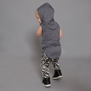 May include: A toddler wearing a gray hooded sleeveless shirt and black and white patterned leggings. The toddler is standing on a gray background.