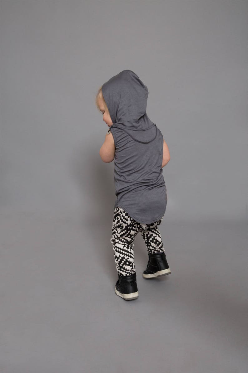 May include: A toddler wearing a gray hooded sleeveless shirt and black and white patterned leggings. The toddler is standing on a gray background.