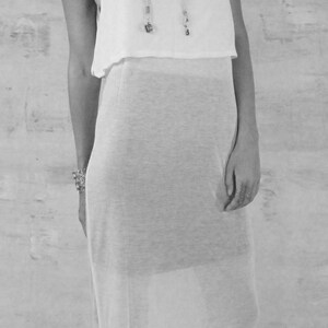 May include: A woman wearing a white sleeveless maxi dress with a sheer overlay. She is wearing black platform sandals and a beaded necklace.
