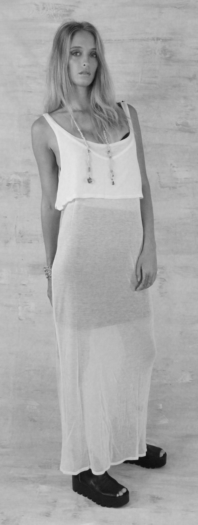 May include: A woman wearing a white sleeveless maxi dress with a sheer overlay. She is wearing black platform sandals and a beaded necklace.