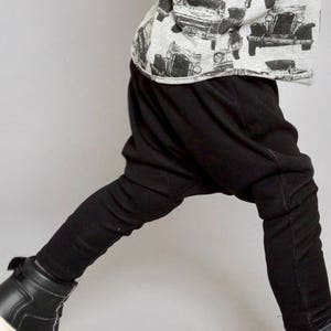 May include: A child wearing black harem pants and black high-top sneakers. The child is wearing a grey and white shirt with a black and white print of vintage cars.