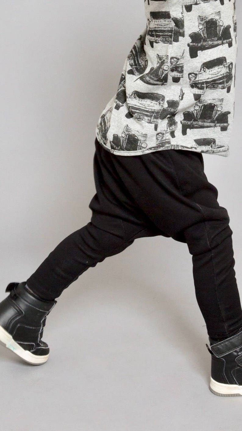 May include: A child wearing black harem pants and black high-top sneakers. The child is wearing a grey and white shirt with a black and white print of vintage cars.