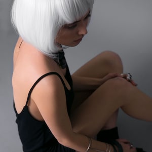 May include: A woman with short, white hair is wearing a black tank top and sitting with her legs crossed. She is wearing a silver bracelet on her wrist and several rings on her fingers.