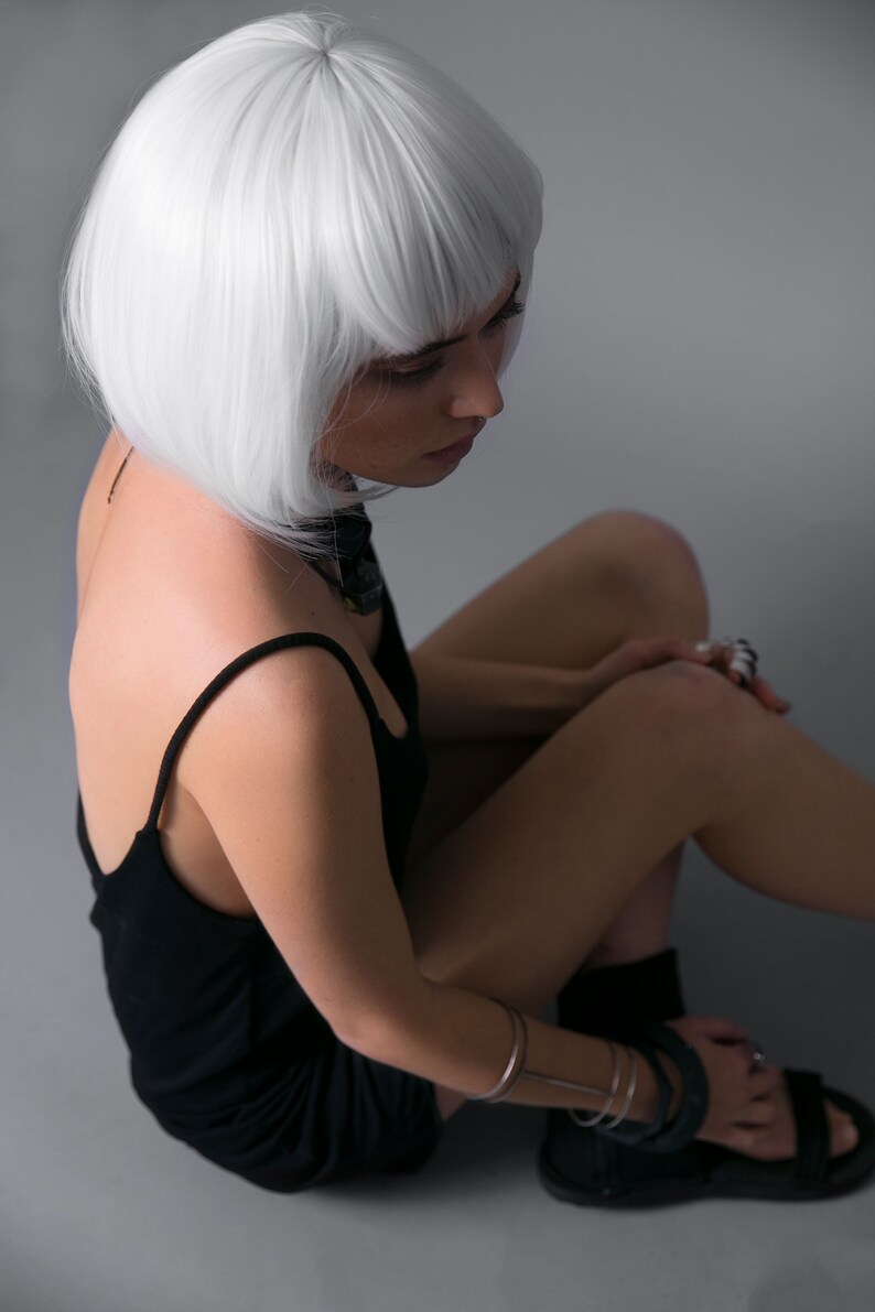 May include: A woman with short, white hair is wearing a black tank top and sitting with her legs crossed. She is wearing a silver bracelet on her wrist and several rings on her fingers.