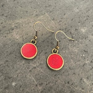 May include: A pair of red and gold dangle earrings with a round, sparkly red centre. The earrings have a simple, minimalist design.