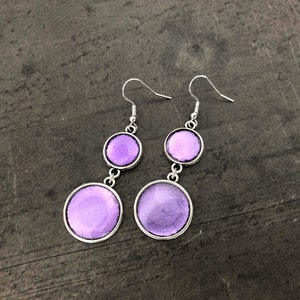 Peut inclure: Une paire de boucles d'oreilles pendantes avec deux cercles en verre violet sur chaque boucle d'oreille. Les boucles d'oreilles ont des crochets argentés.