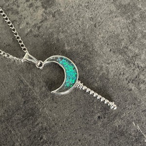 May include: A silver chain necklace with a crescent moon pendant. The moon is filled with green glitter.