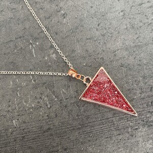 Könnte beinhalten: Eine silberne Kettenkette mit einem roségoldenen Dreiecksanhänger, der mit rotem Glitzer gefüllt ist.