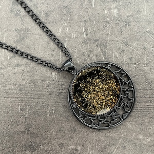 May include: A black chain necklace with a round pendant featuring a black and gold glitter design surrounded by a star-shaped frame.