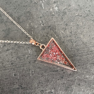 Könnte beinhalten: Eine Halskette aus Roségold mit einem dreieckigen Anhänger, der mit rotem Glitzer gefüllt ist.