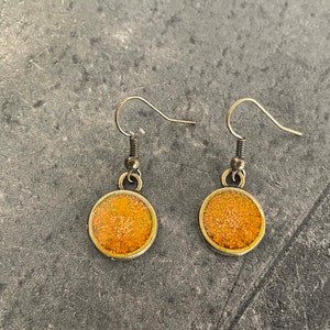 May include: A pair of gold-toned earrings with round orange glitter accents. The earrings have a simple hook design.