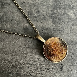Könnte beinhalten: Eine goldene Halskette mit einem runden Anhänger mit einem braunen und goldenen Glitzerdesign.