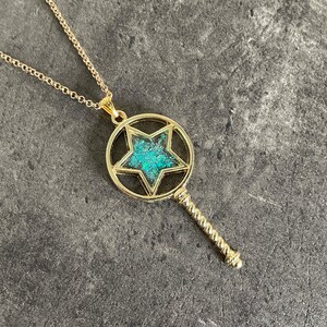 May include: A gold necklace with a pendant shaped like a key. The key has a circular head with a five-pointed star inside. The star is filled with a turquoise-colored material.