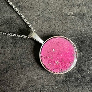 Könnte beinhalten: Eine silberne Kettenkette mit einem runden Anhänger, der mit pinkfarbenem Glitzer gefüllt ist, der in klarem Harz eingebettet ist.