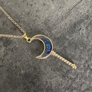 May include: A gold necklace with a crescent moon pendant filled with blue glitter. The pendant has a twisted gold stem.