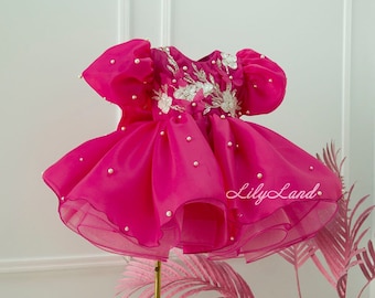 Puede incluir: Un vestido fucsia vibrante con una falda en capas y mangas abullonadas. El corpiño está adornado con adornos florales plateados y perlas dispersas. El vestido se exhibe sobre un soporte dorado, con el texto "Lily Land" visible.