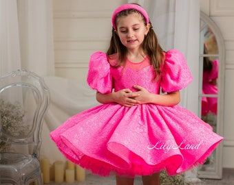 Puede incluir: Una niña pequeña con un vestido de tul rosa brillante con mangas abullonadas y una diadema. El vestido tiene una textura brillante y una falda de capas. La niña está de pie sobre un suelo de madera con una silla transparente en el fondo.