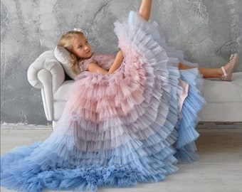 Peut inclure: Une jeune fille portant une robe en tulle à plusieurs couches avec un dégradé de rose, bleu et bleu clair. La robe a un ourlet à volants et la fille est assise sur un canapé blanc, la jambe levée.