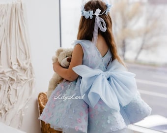Puede incluir: Una niña pequeña con un vestido azul claro con un gran lazo en la espalda. El vestido está decorado con pequeñas flores y la niña lleva un oso de peluche. La niña lleva una corona de flores y mira por una ventana.