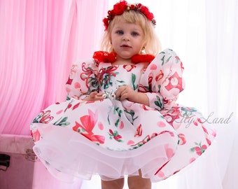Puede incluir: Una niña pequeña con un vestido blanco con estampado floral rojo y verde y un lazo rojo. Lleva una corona de flores rojas y zapatos rojos. El vestido tiene una enagua de tul blanco. El fondo es una cortina rosa.
