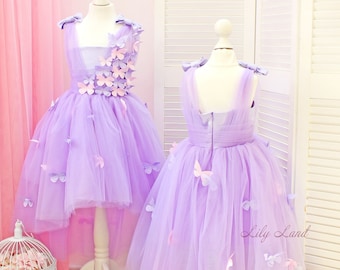 May include: Two lilac tulle dresses with butterfly embellishments. The dress on the left has a high-low hemline. The dress on the right has a full skirt. Both dresses have a sleeveless design and a V-neckline. The dresses are displayed on mannequins against a white slatted background. The text "Lily Land" is visible in the lower right corner of the image.