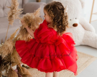 May include: A little girl wearing a red tulle dress with multiple layers of ruffles. The dress has a sweetheart neckline and is sleeveless. The girl is standing on a woven rug and is looking to the side.