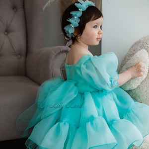 May include: A young child wearing a turquoise off-the-shoulder dress with ruffled layers and puffy sleeves. The dress has the text "Lily Land" on it. The child is also wearing a matching floral headband and gold glitter shoes.