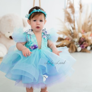 May include: A young girl wearing a light blue tulle dress with floral embellishments and a blue flower crown. The dress has a layered skirt and a V-neckline. The girl is wearing pink shoes with white pom-poms. The text "Lily Land" is visible in the image.