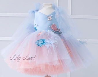 May include: A light blue tulle dress with a pink tulle underskirt. The dress is decorated with embroidered sea creatures, pearls, and feathers. The dress is on a white dress form. The text "Lily Land" is visible on the dress.