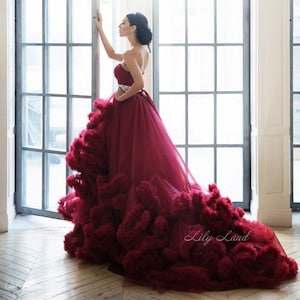 Op de afbeelding: Een vrouw die een strapless, bordeauxrode tulejurk draagt met een gerimpelde rok en een glinsterende riem. De jurk heeft een lange sleep en is erg vol. De vrouw staat voor een raam met een witte achtergrond.